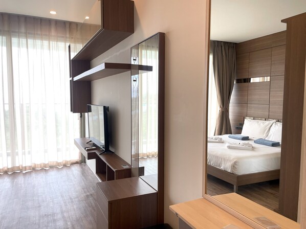 A modern hotel room with a large mirror, a flat-screen TV, a wooden desk, a bed with a headboard, and a window with sheer curtains. - Sea view studio at Oceana by Lofty (Kamala)