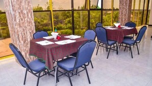 A dining area with tables set for four, blue chairs, and a view of greenery through large windows. - Muhako Hotel (Dar es Salaam)