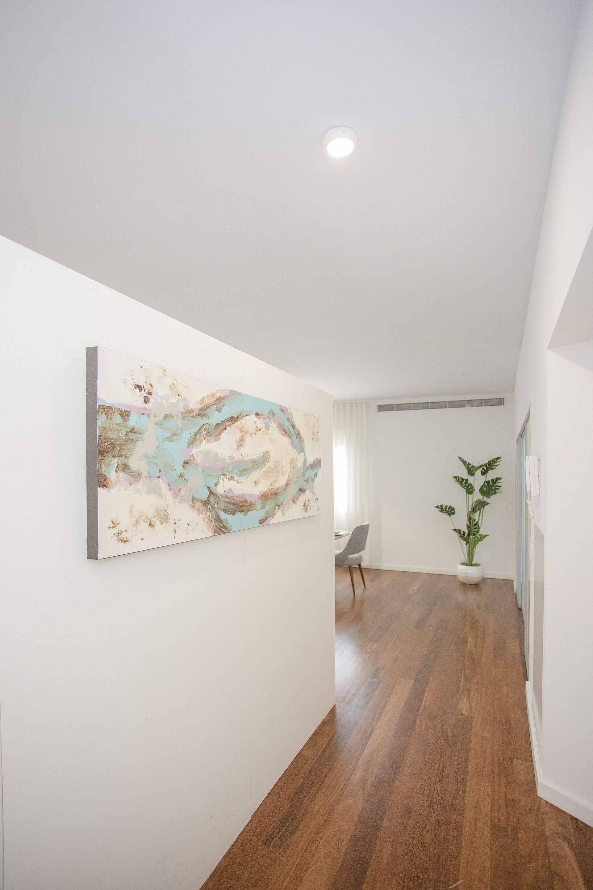 A hallway with a wooden floor, a painting on the wall, a potted plant, and a chair.