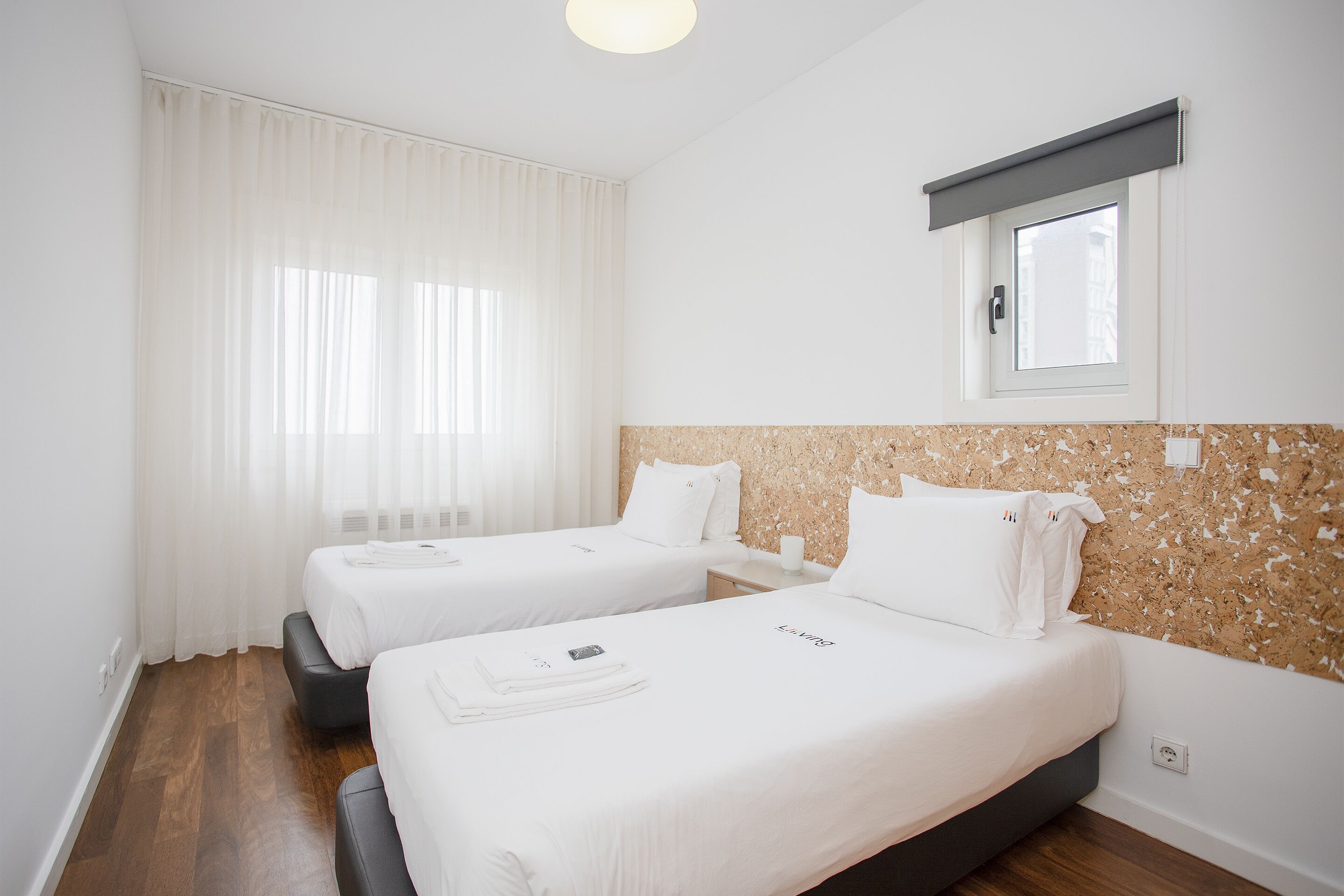 A hotel room with two beds, a window with a view, a wooden floor, and a wall with a textured pattern.