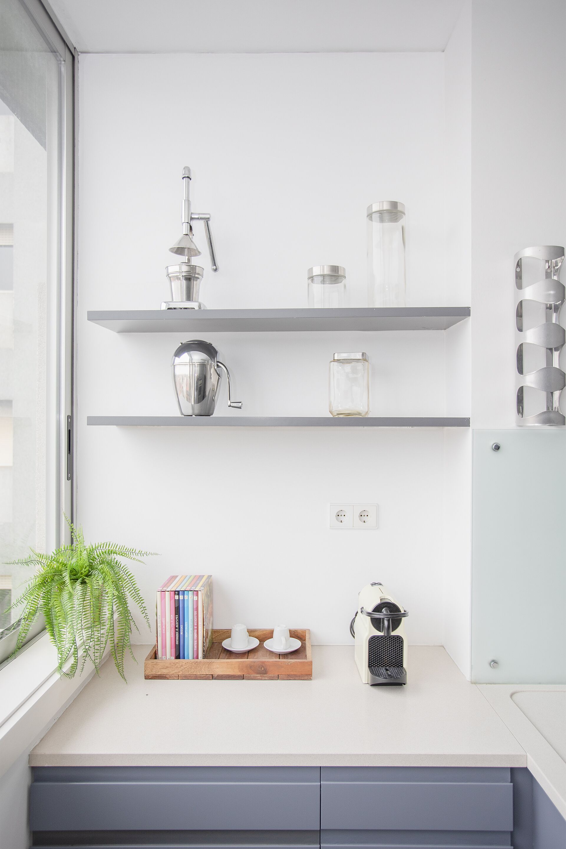 A modern kitchen with white walls, grey shelves, and a window. There are books, a plant, and kitchen appliances on the counter.