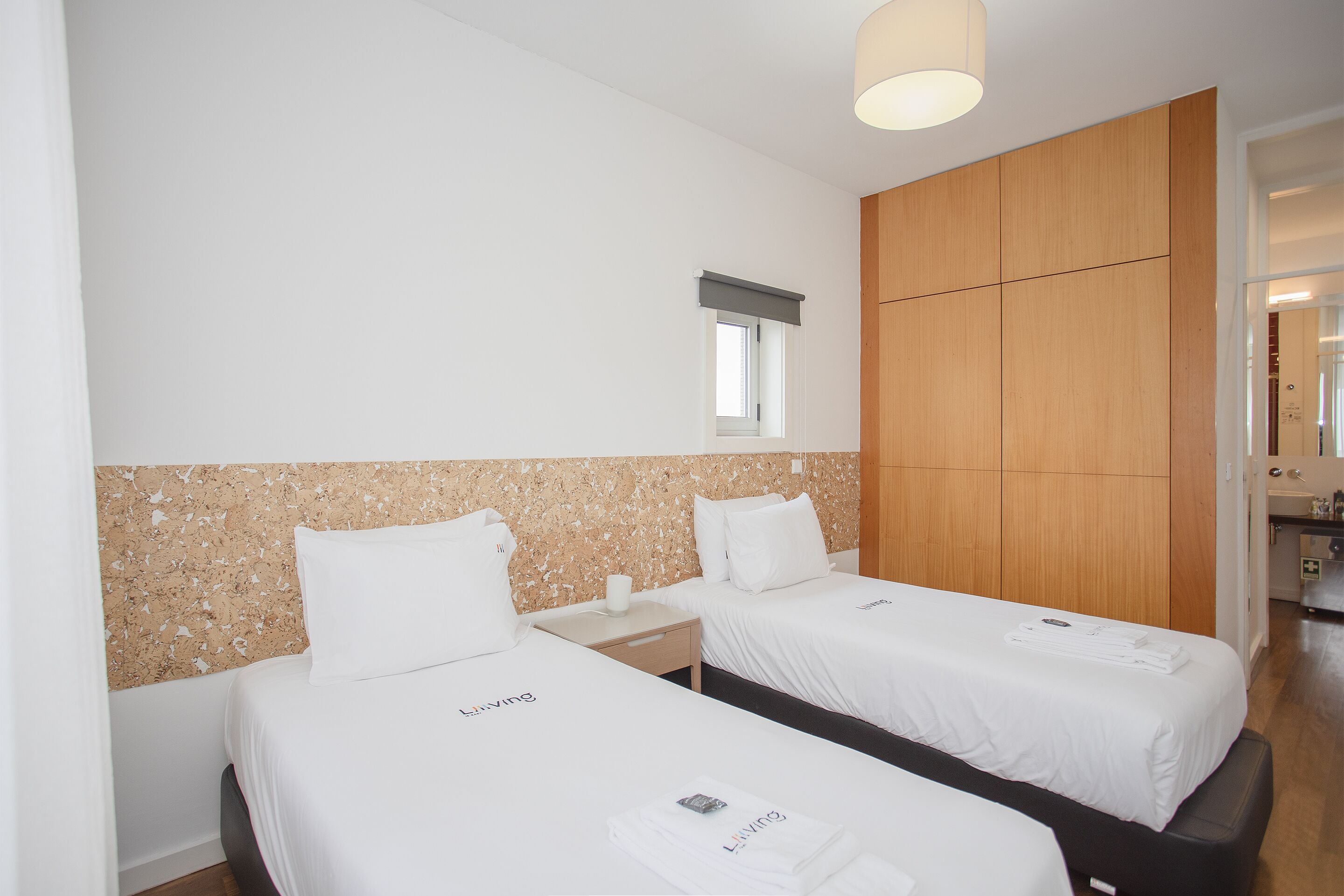 A hotel room with two beds, a wooden wardrobe, a small window, and a bathroom visible through an open door.