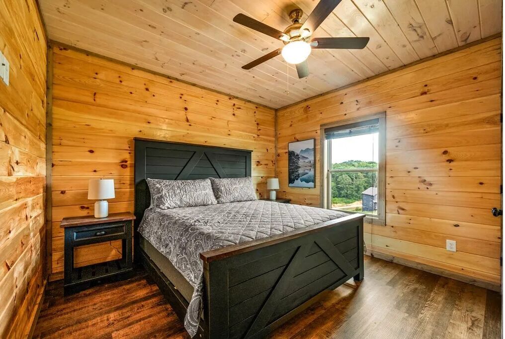 A wooden-paneled bedroom with a bed, two bedside tables, a ceiling fan, and a window with a view of greenery.