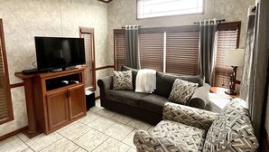 Una sala de estar con un ventilador de techo, un sofá, un sillón estampado, un televisor sobre un mueble de madera y una ventana con persianas.