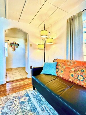 Una sala de estar con un sofá de cuero negro, un cojín estampado, una alfombra azul y una lámpara de pie.