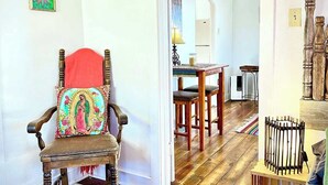 Una habitación con piso de madera, una silla de madera con un cojín colorido, una mesita con una lámpara y una cama con cabecera de madera.