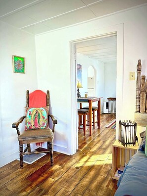 Una habitación con piso de madera, una silla de madera con un cojín colorido, una mesita con una lámpara y una cama con cabecera de madera.
