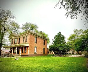 A two-story brick house with a front porch, a well-maintained lawn, and surrounding trees. - La Casita | Southwest Studio Near Downtown Raton (Raton)