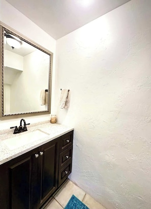 A bathroom with a large mirror, a sink, dark cabinets, and a blue rug on the floor.