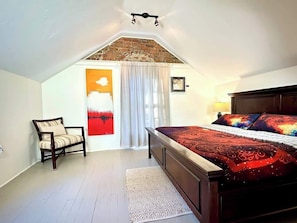 A bedroom with a sloped ceiling, a bed with a patterned bedspread, a chair, a painting, and a window with curtains.