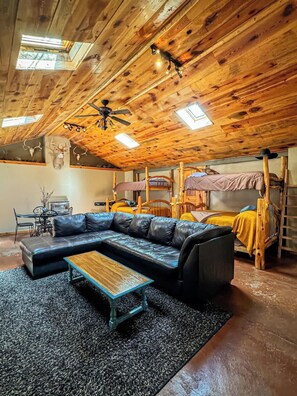 Interior rústico de una cabaña con techo de madera, un sofá de cuero negro, una mesa de centro de madera y una cama litera con una manta amarilla.