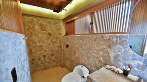 A bathroom with a stone wall, a white toilet, and a wooden shelf above the toilet. - Utopia Tiny Beach House

 (cozoaltepec tonameca)