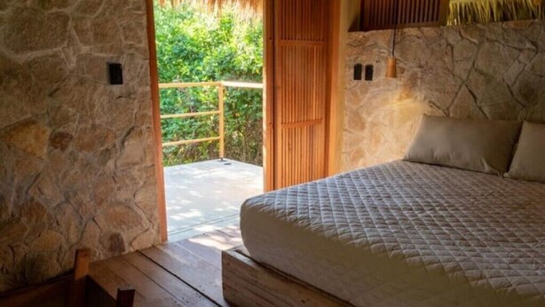 A bedroom with a stone wall, a wooden bed, and a view of greenery outside. - Utopia Tiny Beach House

 (cozoaltepec tonameca)