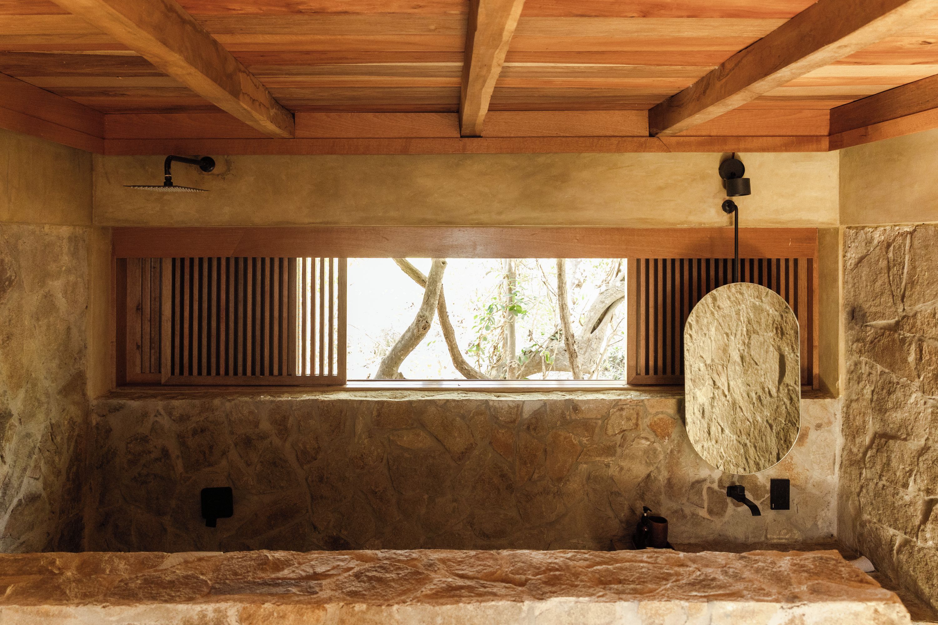 A stone-walled bathroom with a wooden ceiling, a window with vertical blinds, and a stone basin.