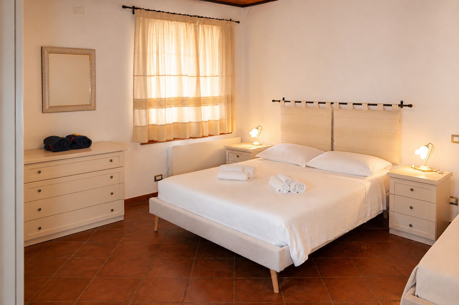Une chambre à coucher comprenant un lit, deux tables de chevet, une commode, une fenêtre avec des rideaux, un luminaire fixé au mur et un miroir fixé au mur.