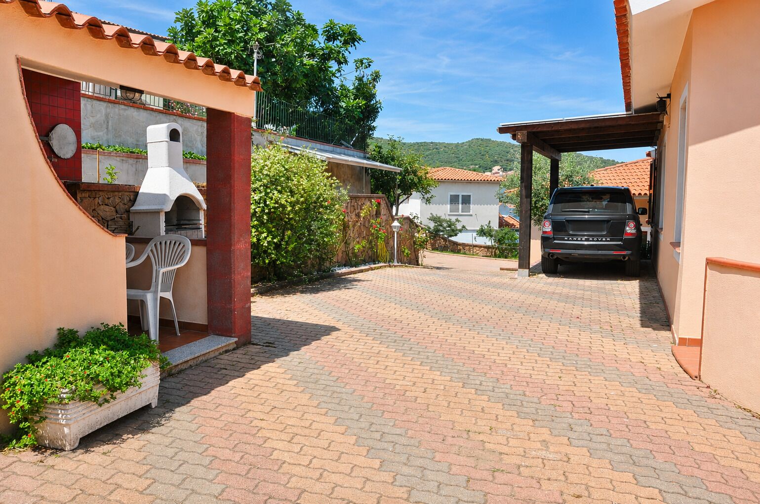 Ein gepflasterter Weg führt zu einem Haus mit Barbereich, überdachter Autogarage und einem darin stehenden Fahrzeug.