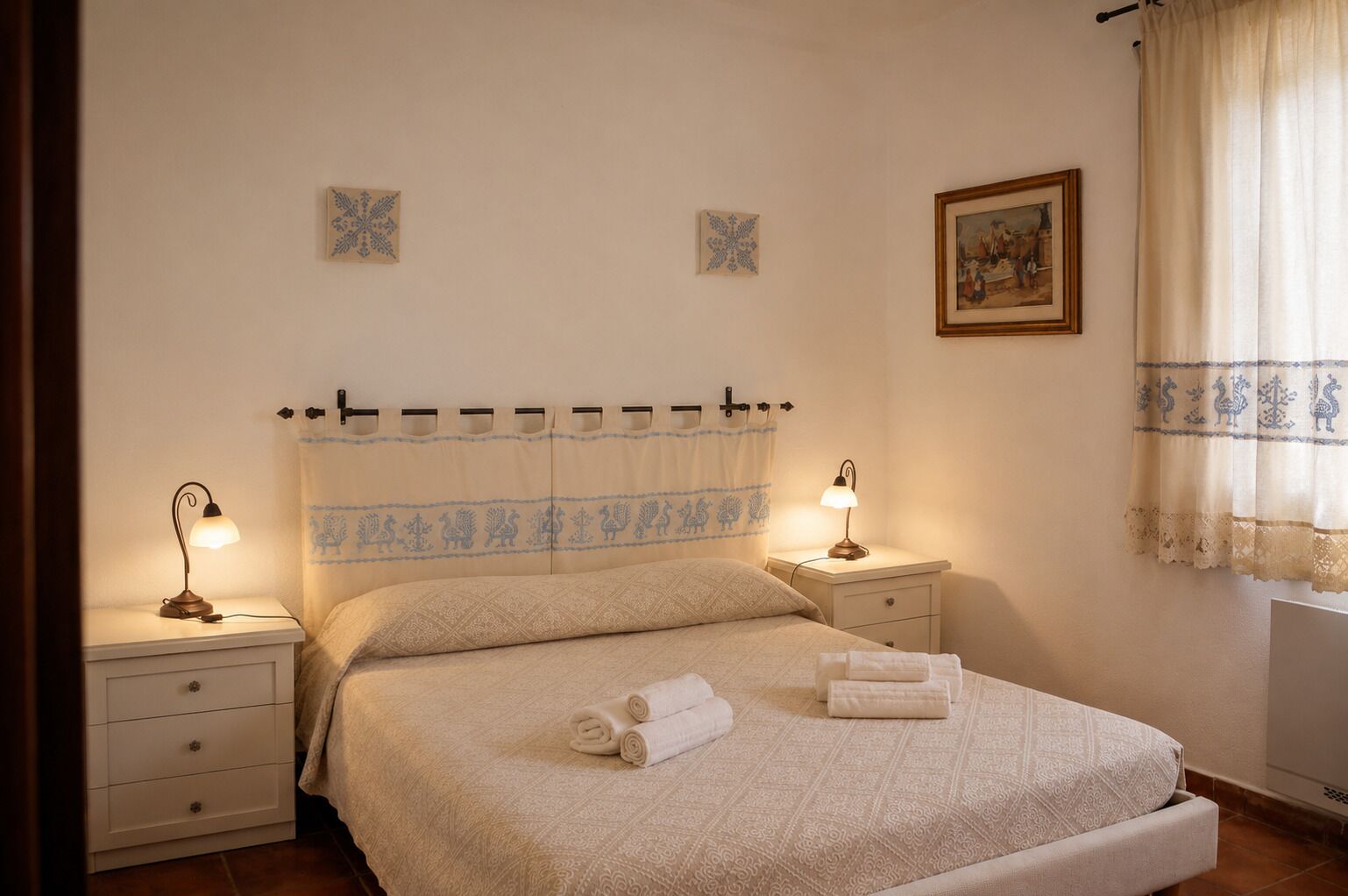 Quarto com uma cama, duas mesas de cabeceira com luminárias, uma janela com cortinas e um quadro na parede.