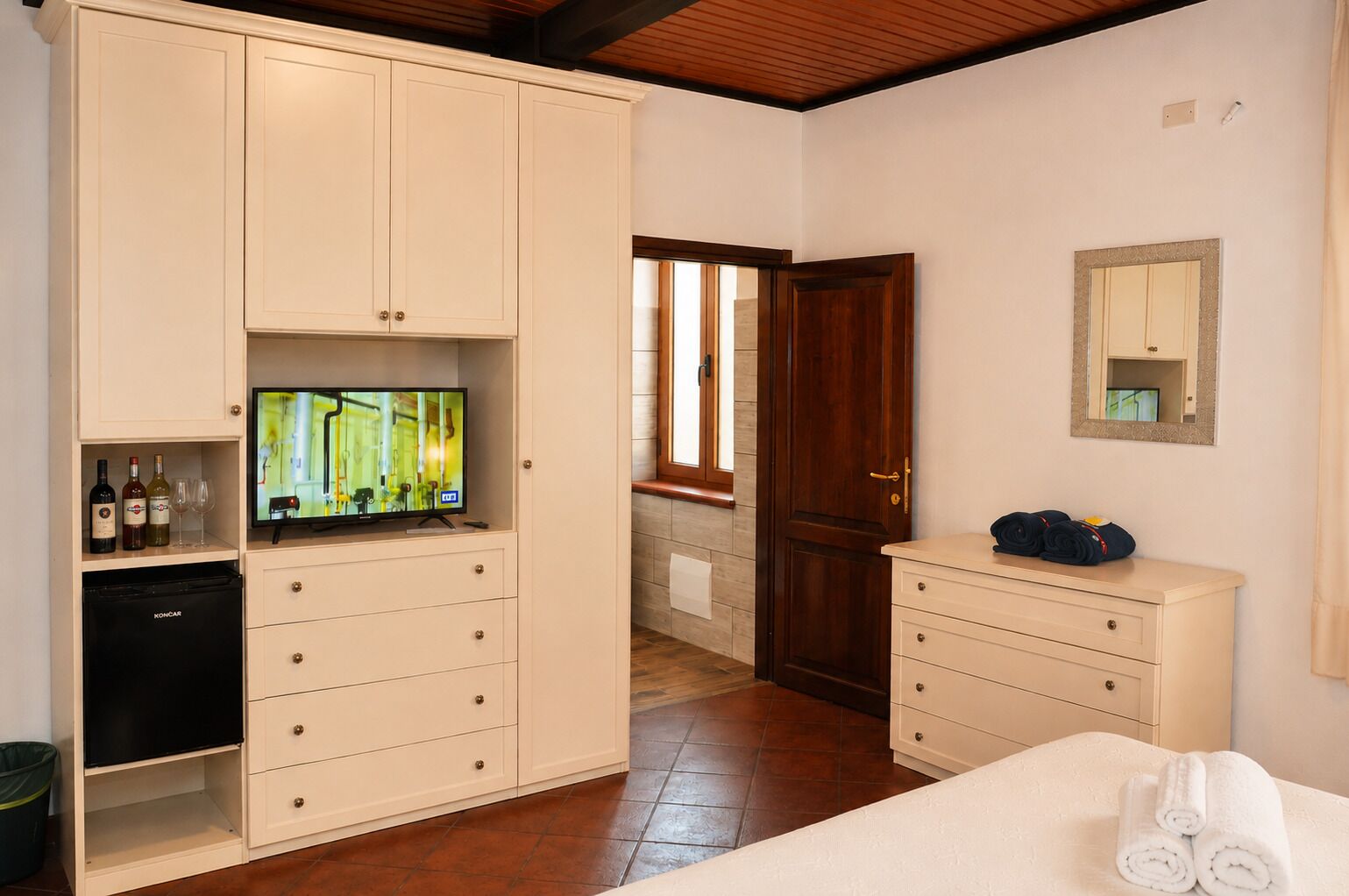 Hotelkamer met een minibar, een televisie, een houten deur, een spiegel en een bed met handdoeken.