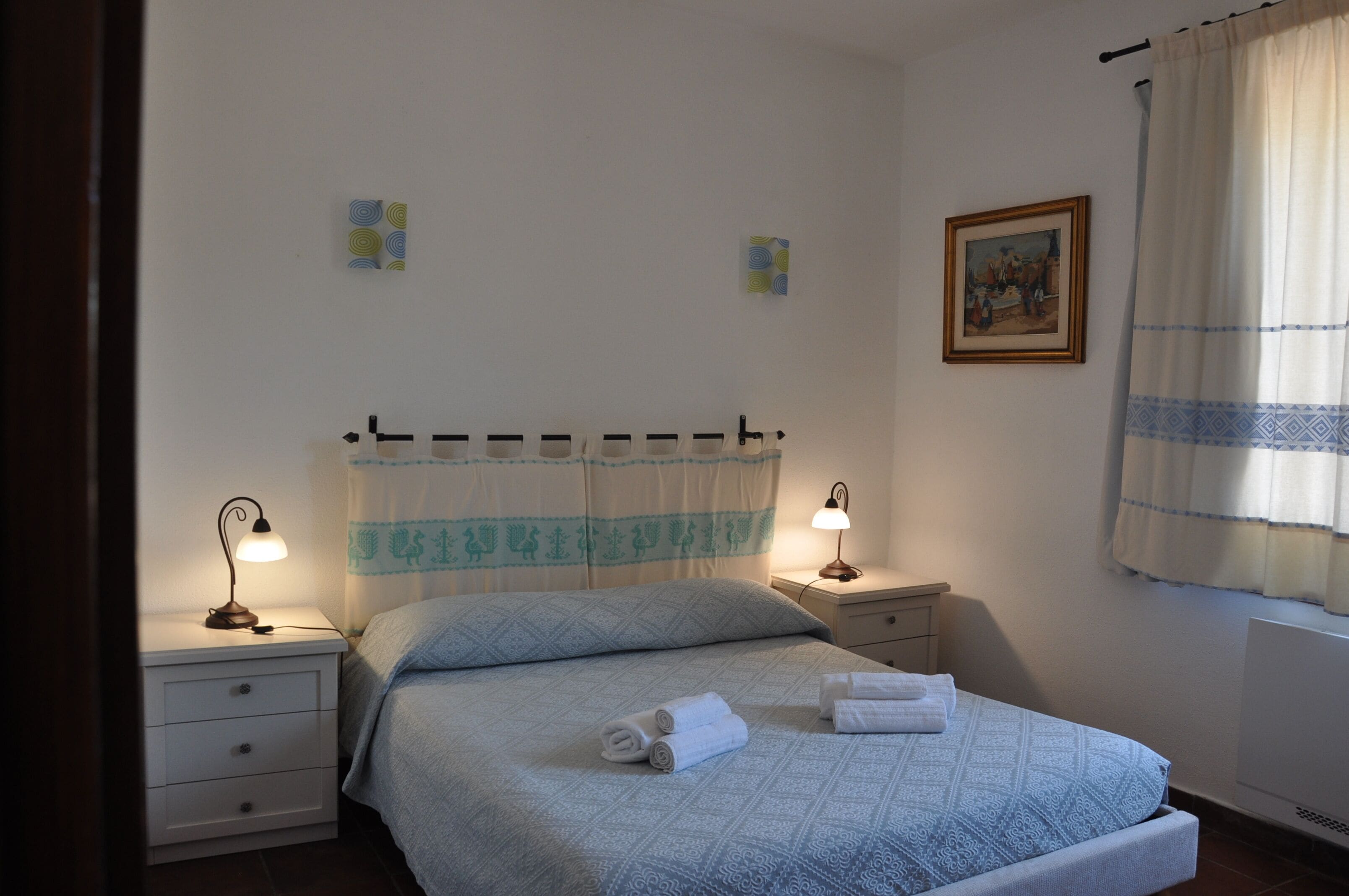 Een slaapkamer met een bed, twee nachtkastjes, een wanddecoratie en een raam met gordijnen.