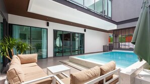 A modern outdoor pool area with a sofa and lounge chairs, glass doors, and a view of the surrounding area. - Family-sized holiday house with private pool, perfect for families and friends! (Chonburi)