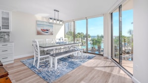 A modern kitchen with white cabinets, a dining area with a glass table and white chairs, and large windows offering a view of the ocean and palm trees. - Beachfront-Luxury Renovated Condo For 13 w/Wrap Balcony @ Edgewater Beach Resort (Panama City Beach)