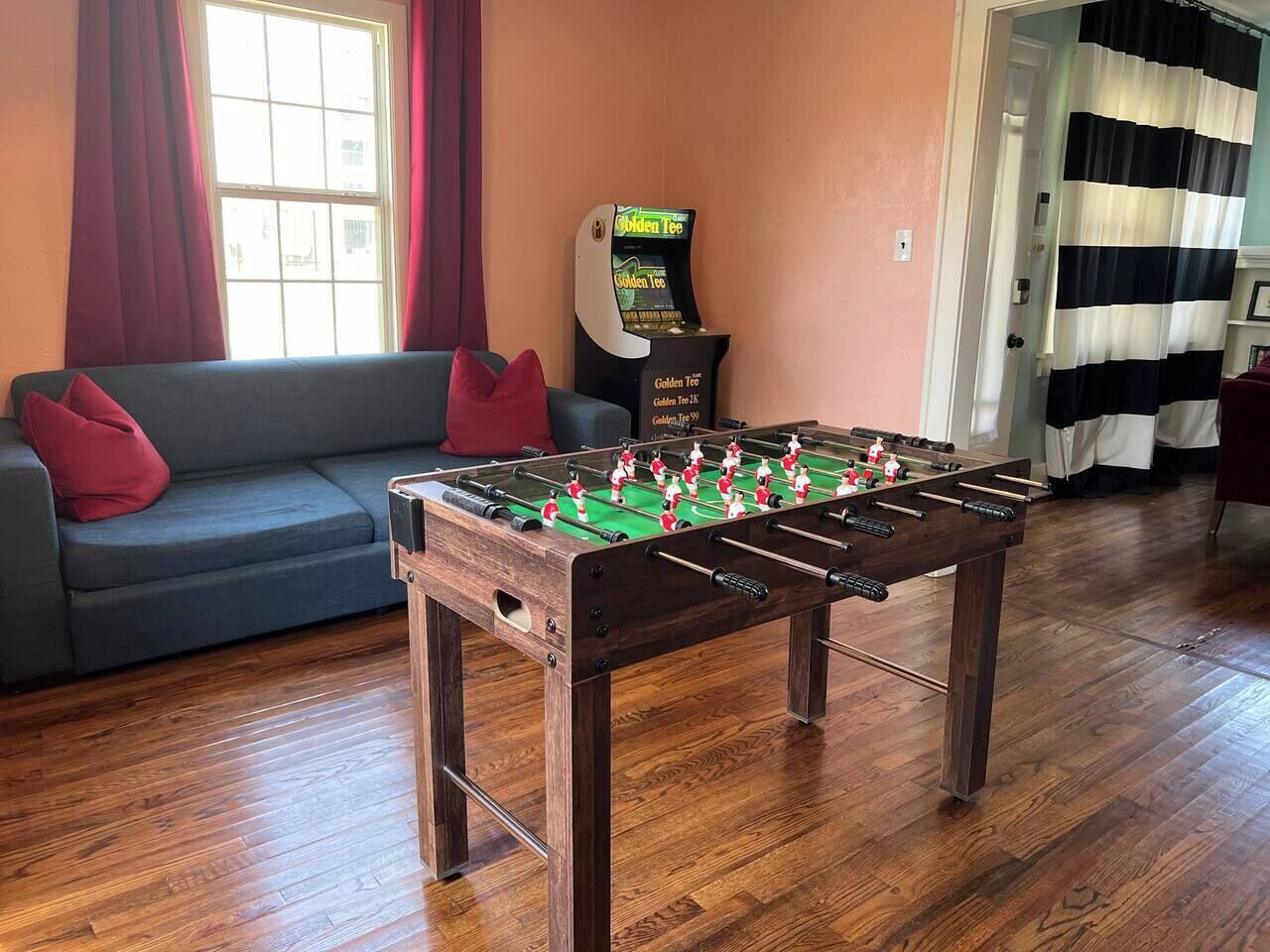 A room with a foosball table, a sofa with red cushions, a window with curtains, and a claw machine.