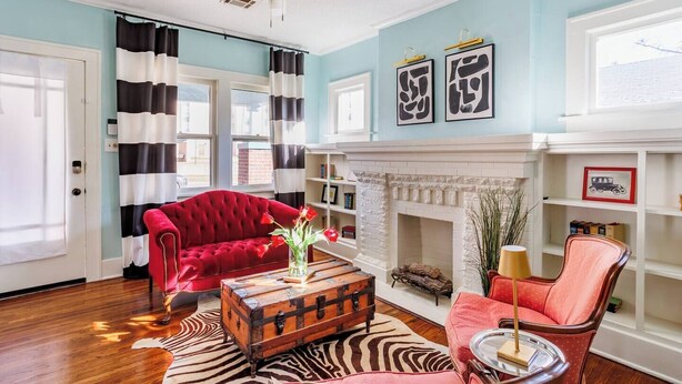 Un salon avec un canapé rouge, une commode en bois, un tapis à motifs zébrés, une cheminée et un ventilateur de plafond.