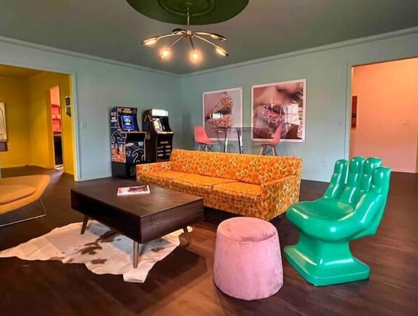 Un salon avec un canapé, une table basse, un pouf rose et un fauteuil vert. On aperçoit des bornes d’arcade à l’arrière-plan.