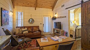 A cozy living room with a brown leather sofa, a wooden coffee table, and a TV on a wooden stand. There is a dining area with a table and chairs, and a kitchen area with a dishwasher. - Vineyard Waterfall Cabin+Free Tasting  on Turkey Creek (Ellijay)