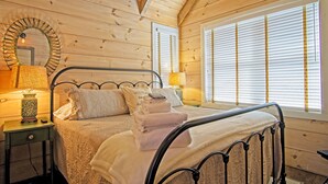 A cozy wooden cabin bedroom with a bed, two bedside lamps, a mirror, and a window with blinds. - Vineyard Waterfall Cabin+Free Tasting  on Turkey Creek (Ellijay)