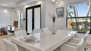 A modern dining area with a white table set for four, a vase with flowers, and a view of palm trees through large windows. - Lynora's Luxe Estate Modern Design (Fort Lauderdale)