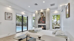 A modern bedroom with a bed, a fireplace, a large window, and a view of a swimming pool. - Lynora's Luxe Estate Modern Design (Fort Lauderdale)