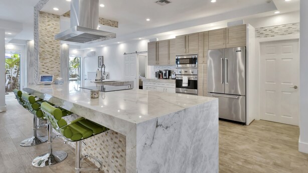 A modern kitchen with stainless steel appliances, a marble island, and a bar area with green stools. - Lynora's Luxe Estate Modern Design (Fort Lauderdale)