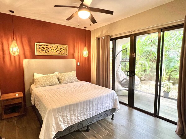 A bedroom with a bed, a ceiling fan, a nightstand, and a view of a garden through a sliding glass door.