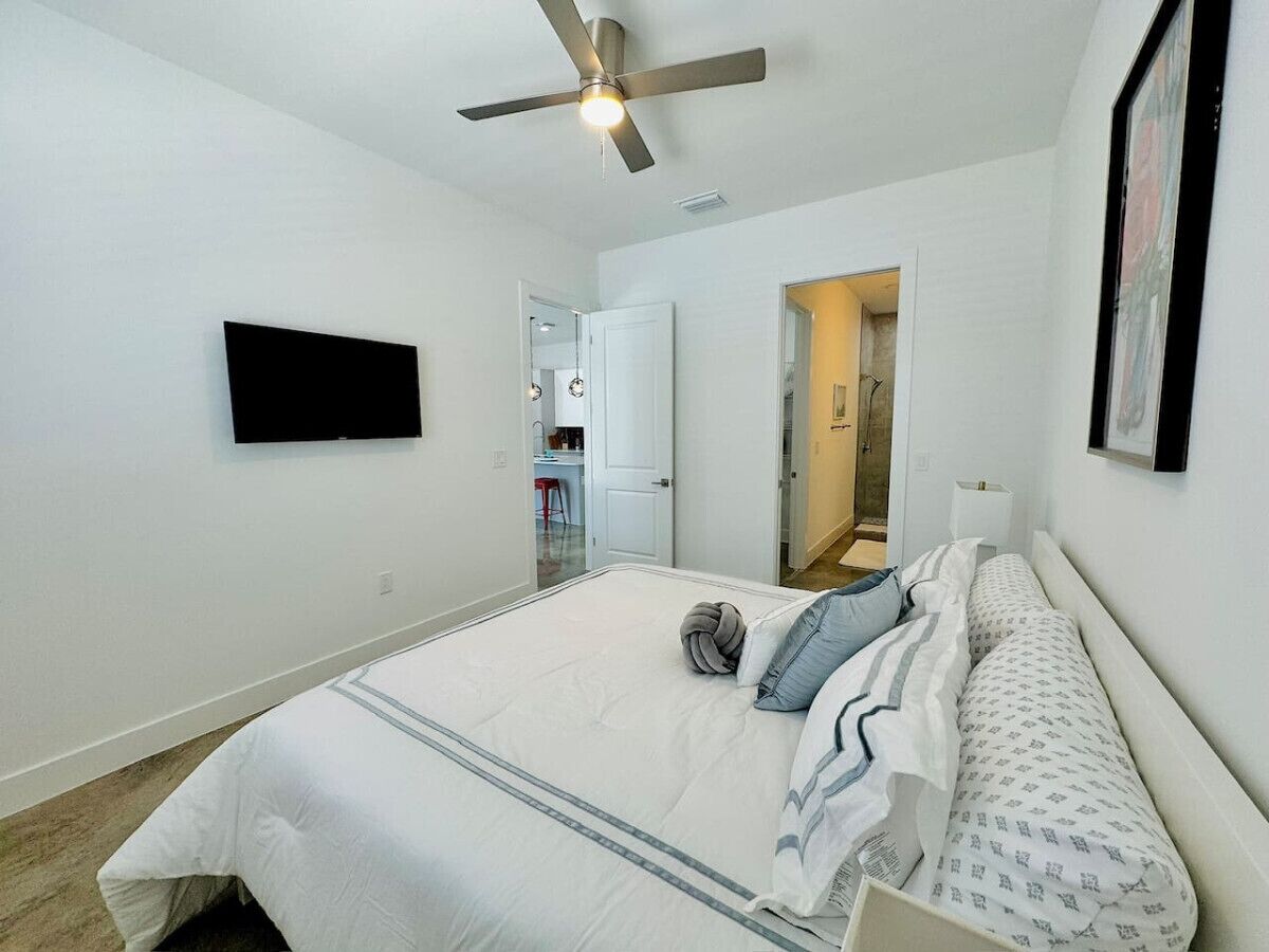 A bedroom with a bed, a ceiling fan, a flat-screen TV, and a door leading to another room.