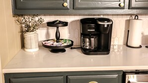 Una barra de cuina amb una cafetera Keurig, un ram de fulles verdes en un vas de vidre blanc i un plat decoratiu amb diversos objectes.