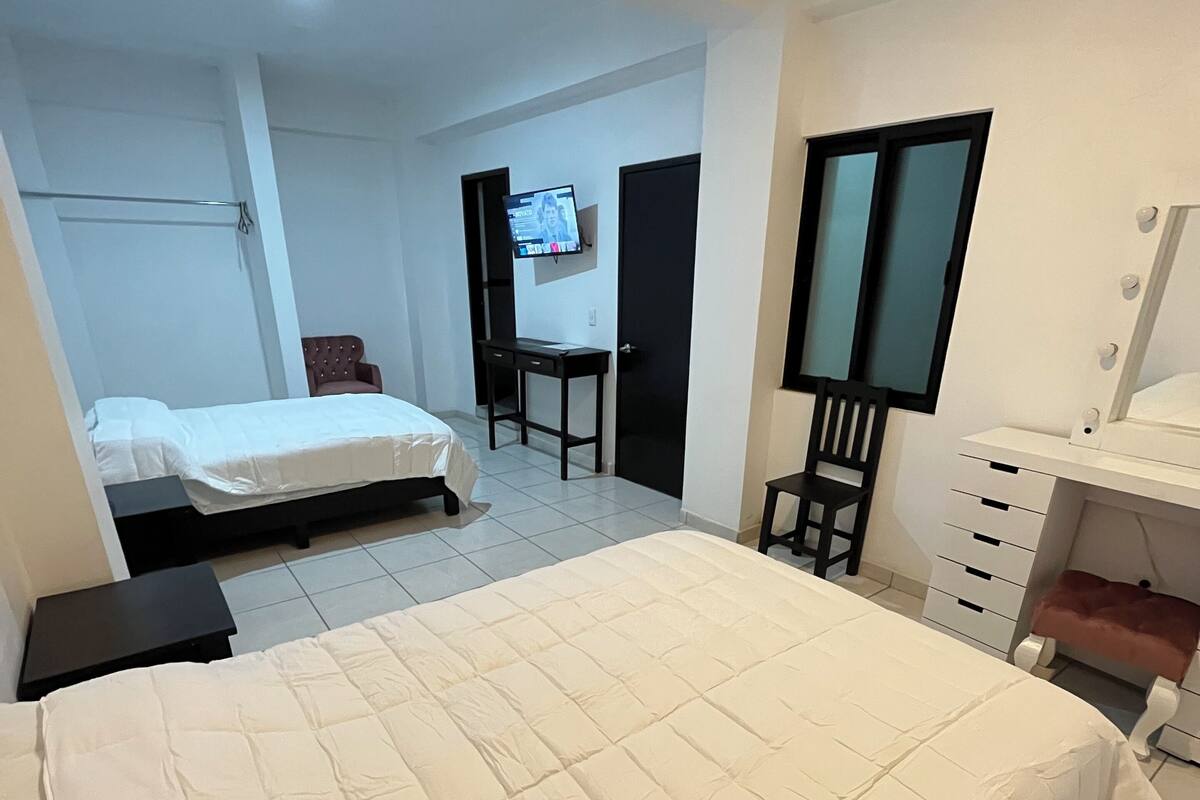 A bedroom with a large bed, a desk, a chair, a vanity with a mirror, and a television on the wall.