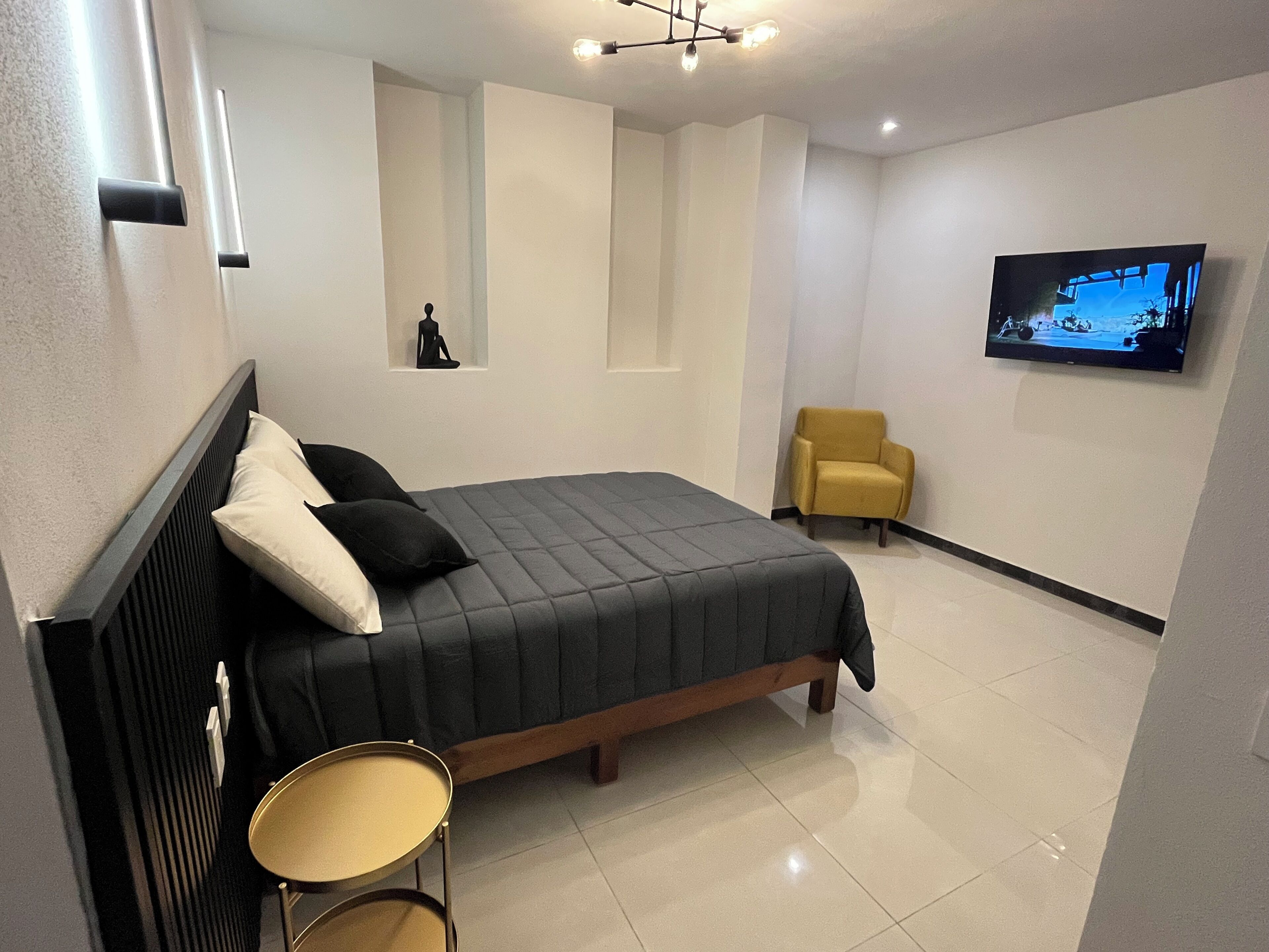 Un dormitorio moderno con una cama, una mesita redonda pequeña, un sillón amarillo y un televisor en la pared.