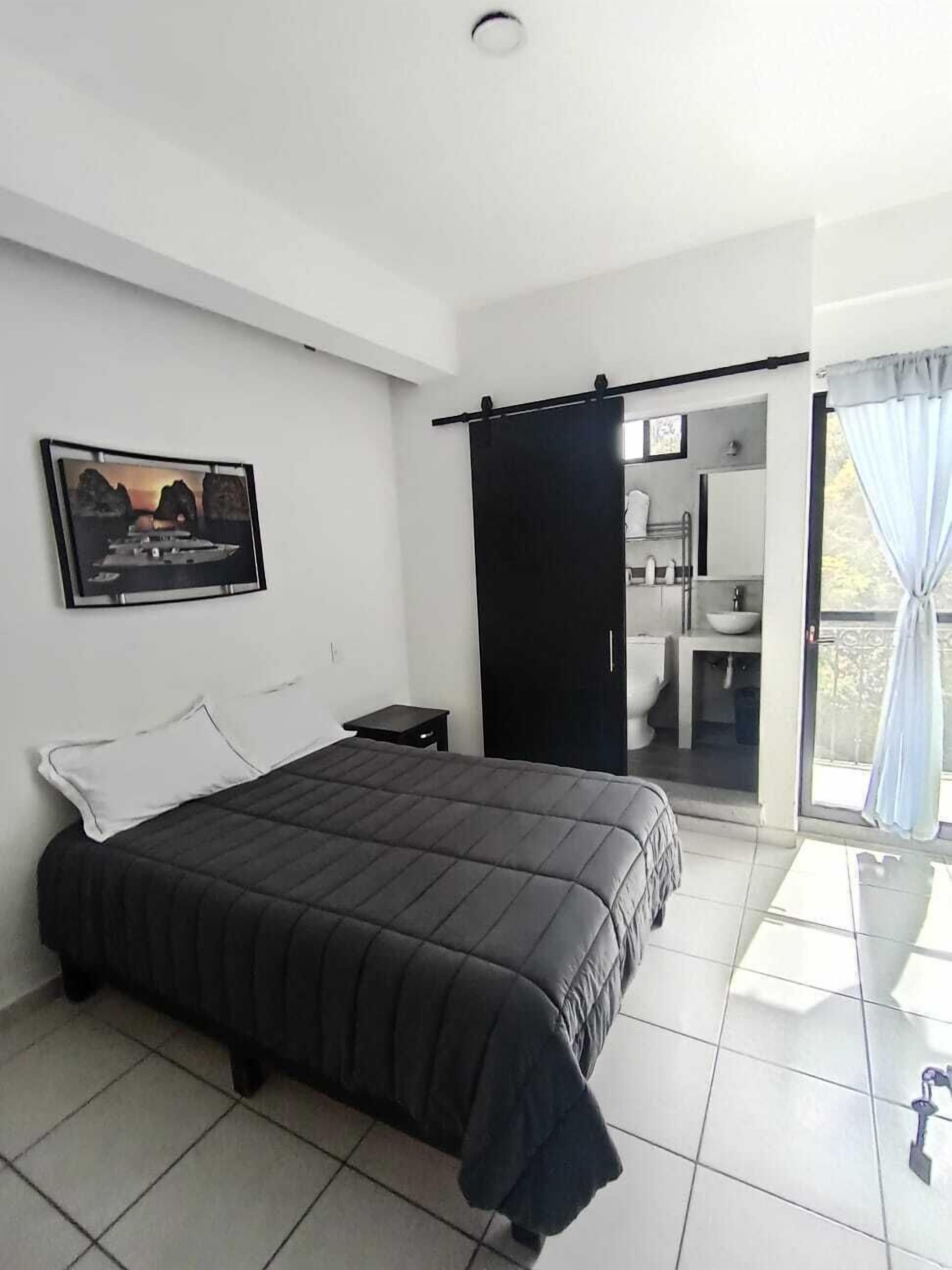 Een moderne slaapkamer met een groot bed, een klein nachtkastje, een zwart gordijn en een zichtbare badkamer door een geopende deur.