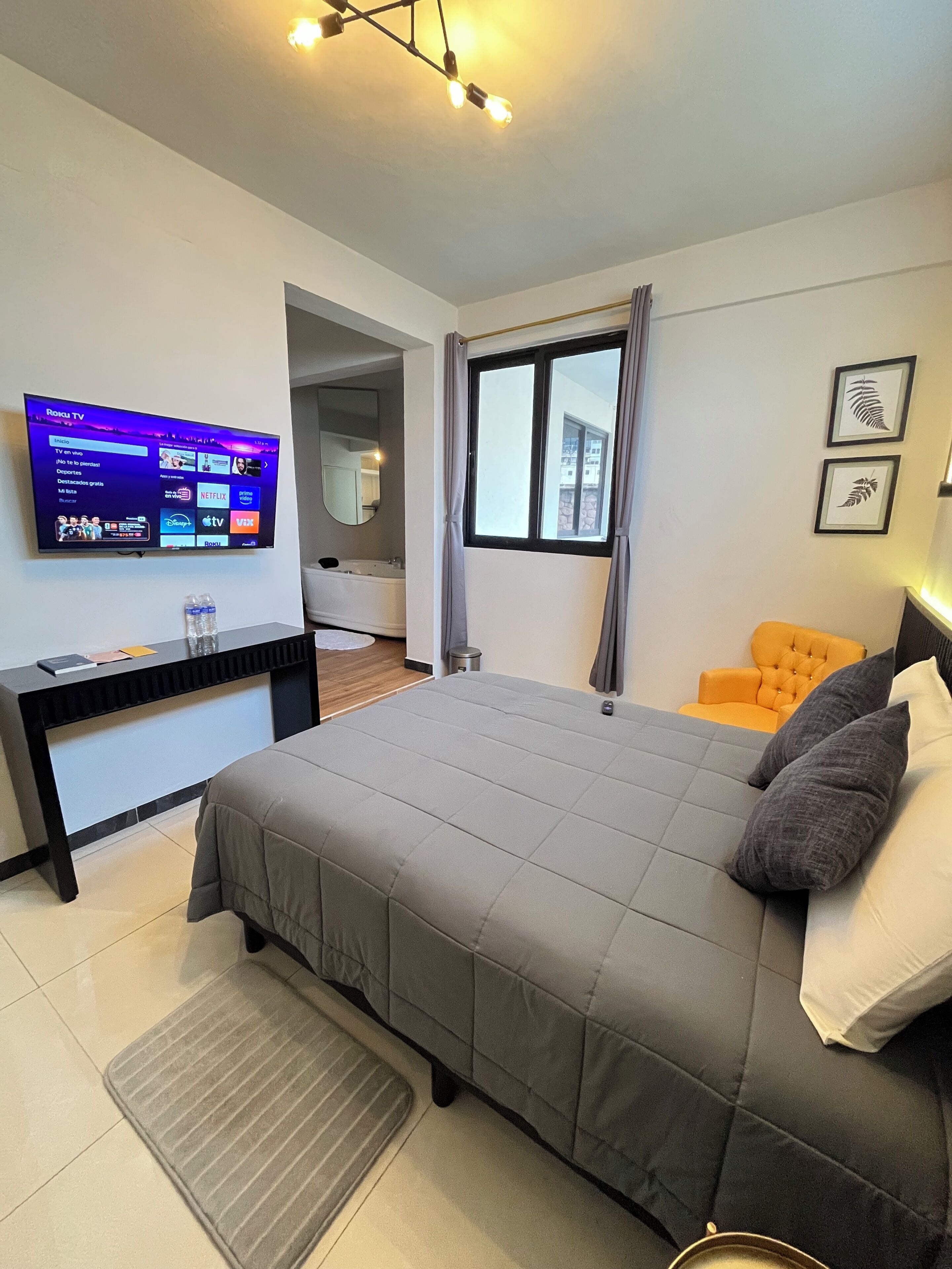Quarto de hotel moderno com cama, escrivaninha, TV exibindo um serviço de streaming, janela com cortinas e banheiro visível por uma porta aberta.
