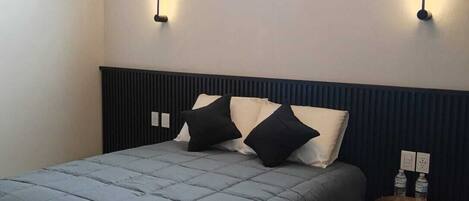 A bedroom with a bed, a window, two wall-mounted lights, a small bedside table with a bottle, and a power outlet.