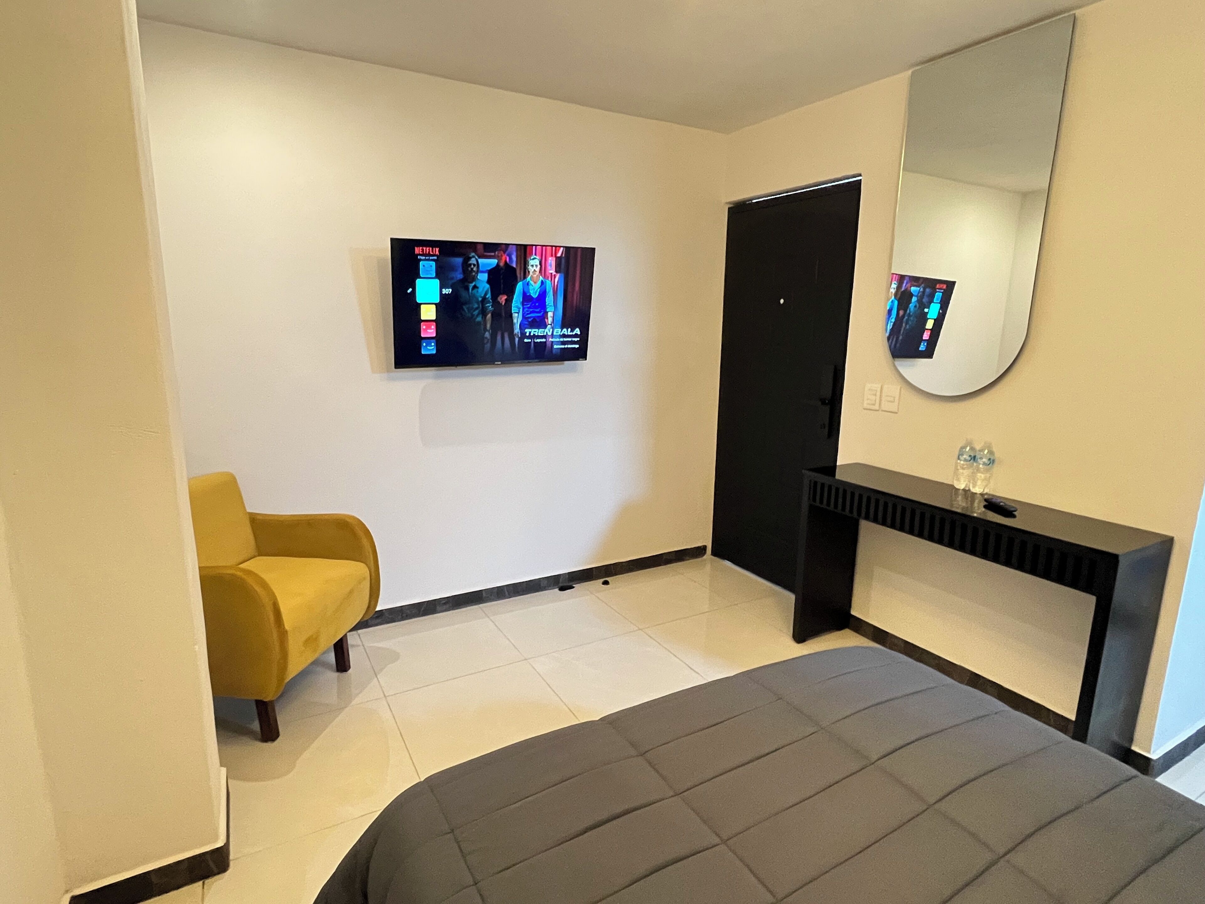 Una habitación de hotel con una cama, un sillón amarillo, una consola negra con un espejo y un televisor que muestra una película.