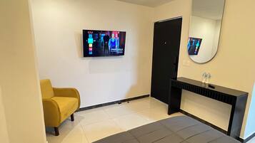 Una habitación de hotel con una cama, un sillón amarillo, una consola negra con un espejo y un televisor que muestra una película.