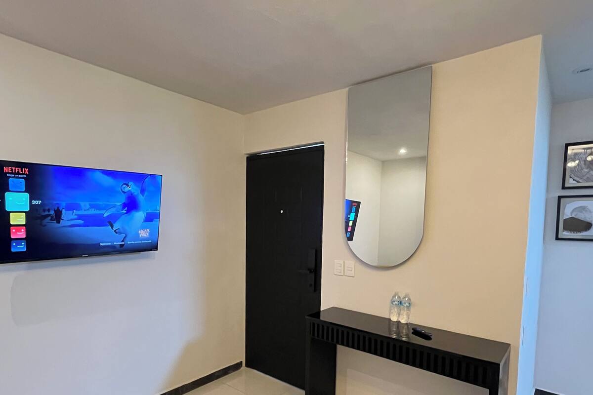 A hotel room with a flat-screen TV mounted on the wall, a black console table, a large mirror, and a bed with a gray headboard.