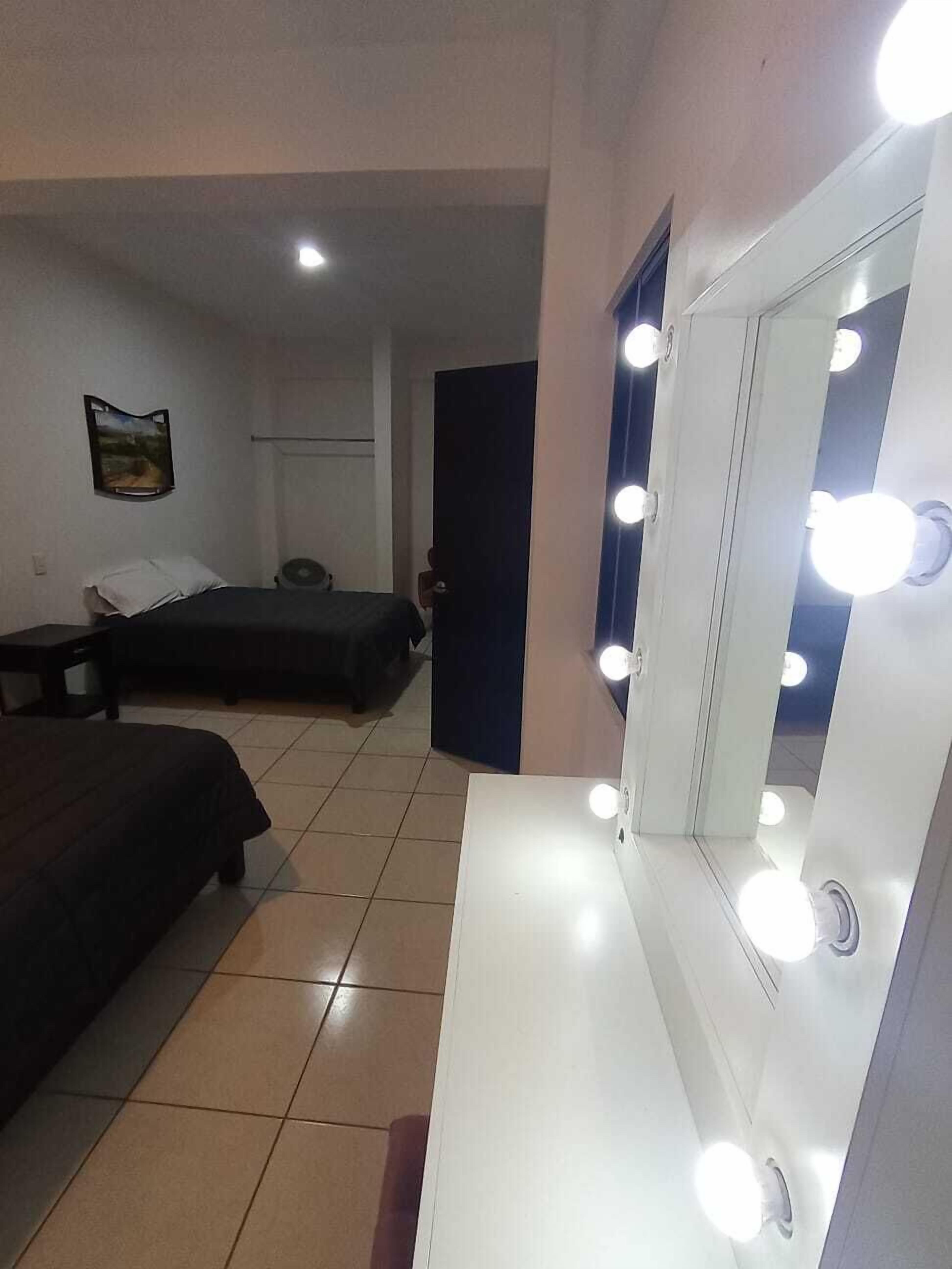 A hotel room with a bed, a bedside table, a mirror with lights, and a door.