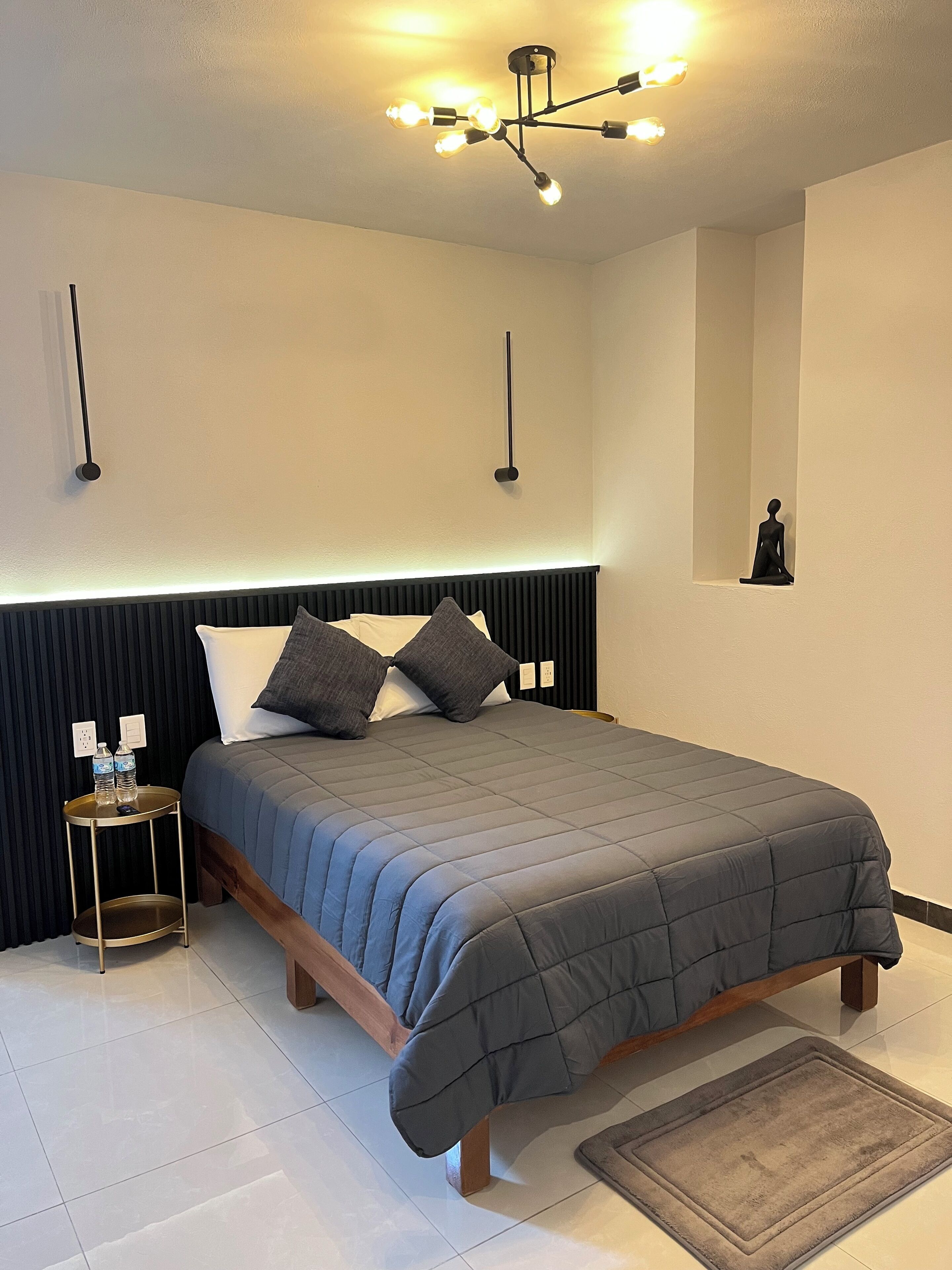 Un dormitorio moderno con cama, mesitas de noche, lámpara de noche y una escultura en la pared, además de un artefacto de iluminación en el techo.