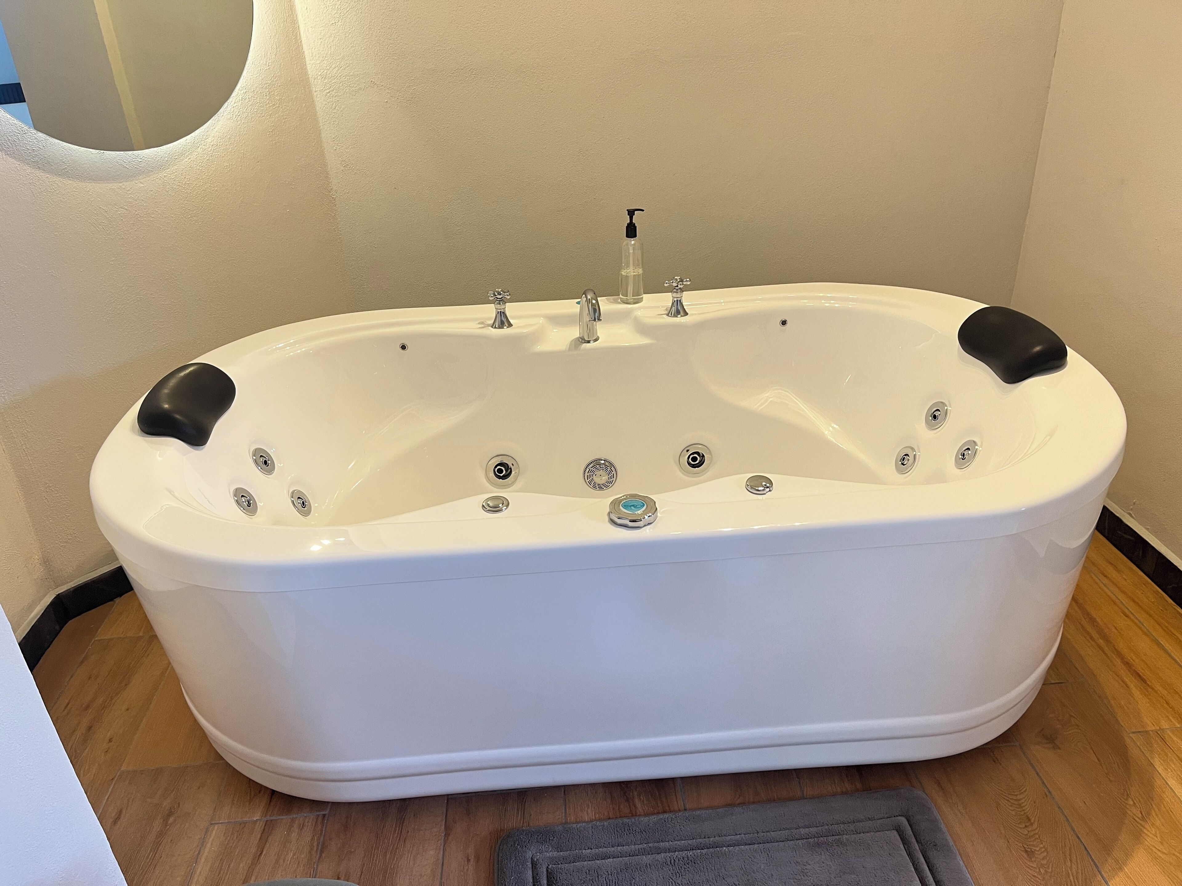 A white freestanding bathtub with multiple faucets and a handheld showerhead, positioned in a corner with a wooden floor and a small rug.