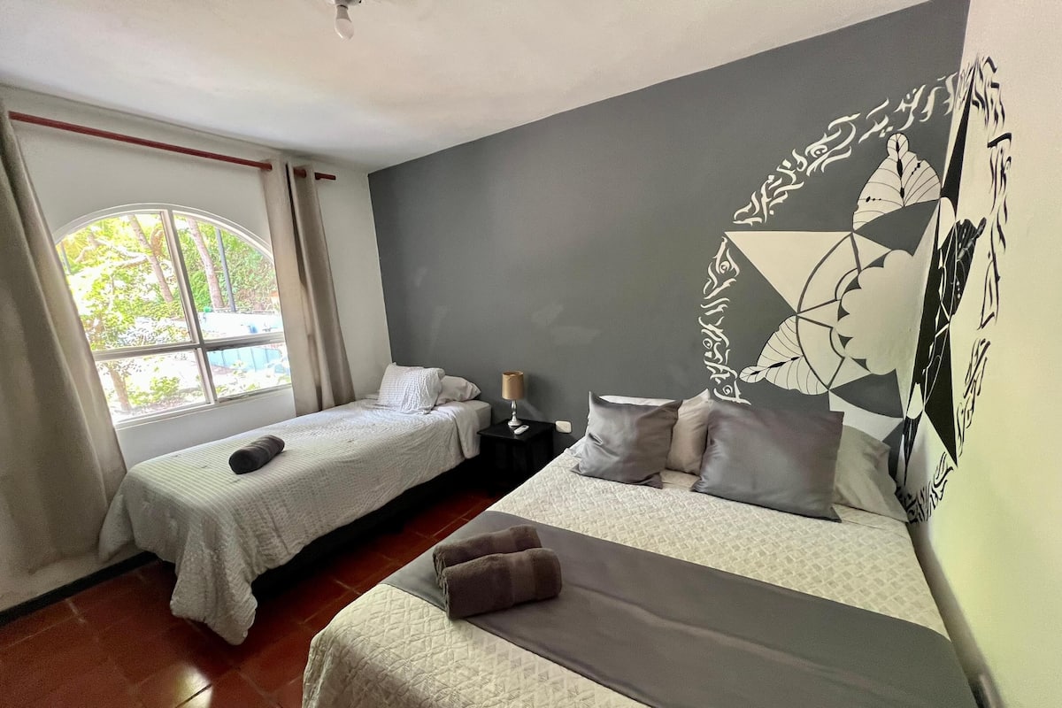 A hotel room with two single beds, a large window with curtains, a wall with geometric patterns, and a ceiling fan.