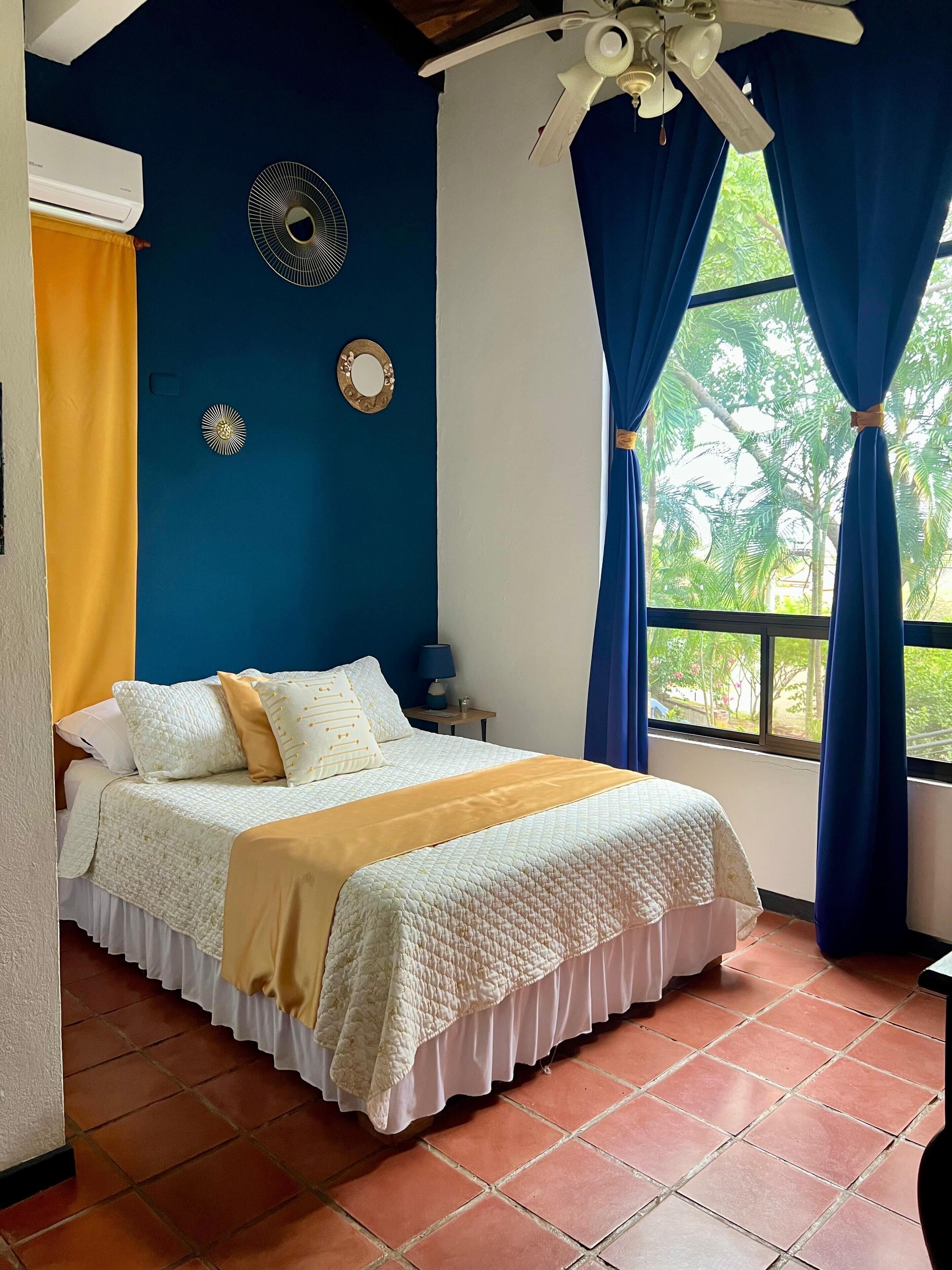 A bedroom with a bed, a ceiling fan, a window with curtains, and a wall-mounted air conditioner.