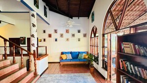 Ein geräumiges Wohnzimmer mit einem blauen Teppich mit Muster, einer Holztreppe, einem blauen Sofa mit Kissen, einem Deckenventilator und einem Bücherregal voller Bücher.